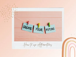 A pastel-themed graphic featuring three light blue cards clipped to a string with colorful clothespins, displaying the words "CREATE YOUR FUTURE" in bold black letters. The background is a soft peach wood panel, and the bottom of the image reads "How to use Affirmations" in elegant cursive. The design includes subtle abstract shapes and a rainbow pattern on the right side